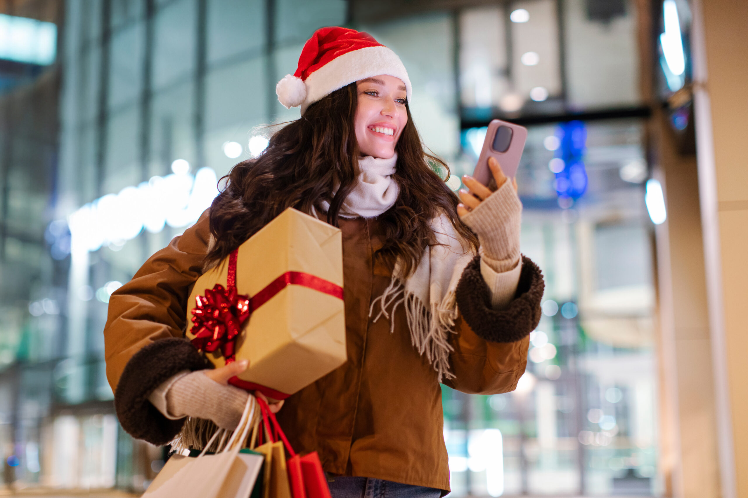 Christmas preparation. Happy lady in Santa hat using cellphone, walking near mall after buying Xmas presents, holding gift box and shopper bags