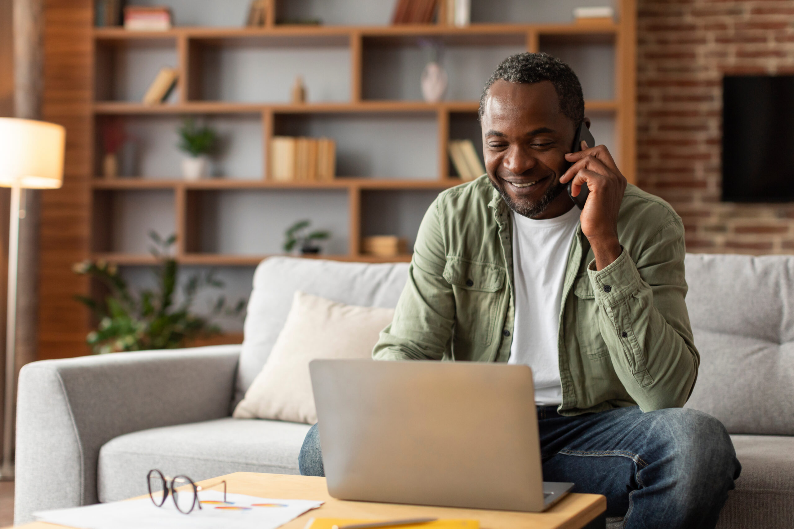 Smiling middle aged black male manager typing on computer, calling by phone, talk with client in living room interior. Work and business at home with modern technology, social distance and new normal